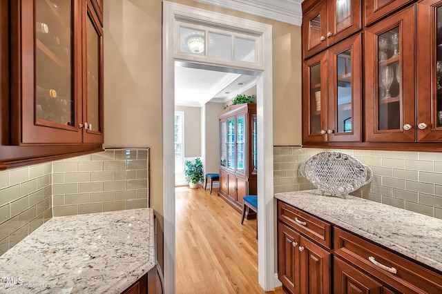 kitchen featuring crown molding, light stone counters, light wood finished floors, backsplash, and glass insert cabinets