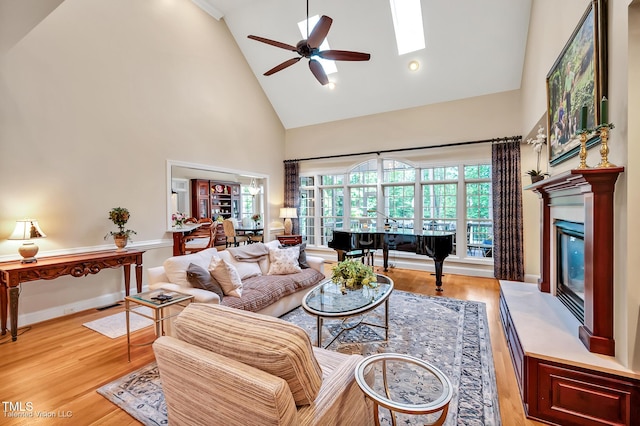 living room featuring high vaulted ceiling, a ceiling fan, light wood-style floors, a glass covered fireplace, and baseboards