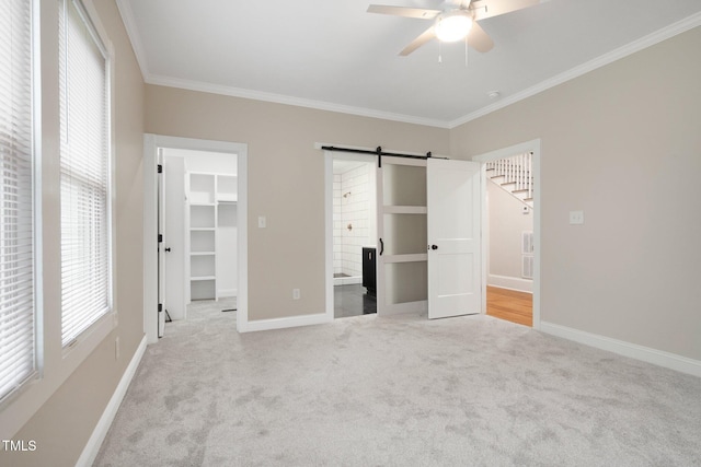 unfurnished bedroom with a barn door, light carpet, a spacious closet, crown molding, and baseboards
