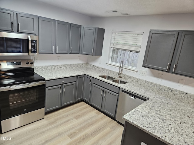 kitchen with appliances with stainless steel finishes, a sink, gray cabinets, light wood-style flooring, and light stone countertops
