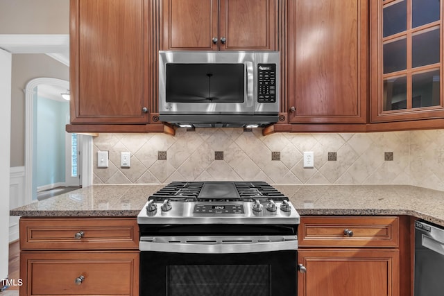 kitchen featuring appliances with stainless steel finishes, glass insert cabinets, light stone countertops, brown cabinetry, and decorative backsplash