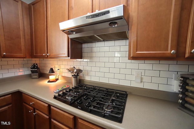kitchen featuring under cabinet range hood, black gas stovetop, backsplash, and brown cabinets
