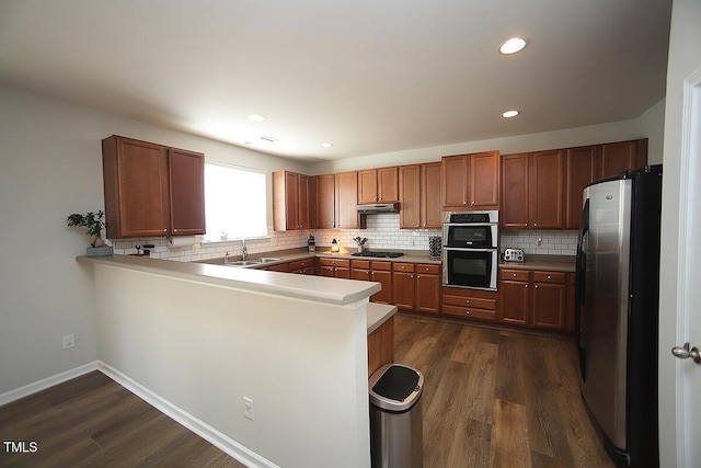 kitchen featuring stainless steel appliances, a sink, a peninsula, decorative backsplash, and dark wood-type flooring