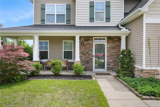 entrance to property featuring covered porch, stone siding, a shingled roof, and a lawn