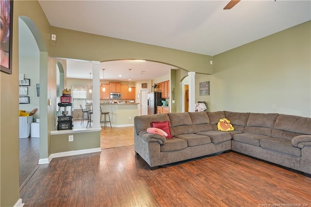 living area featuring wood finished floors, arched walkways, decorative columns, a ceiling fan, and baseboards