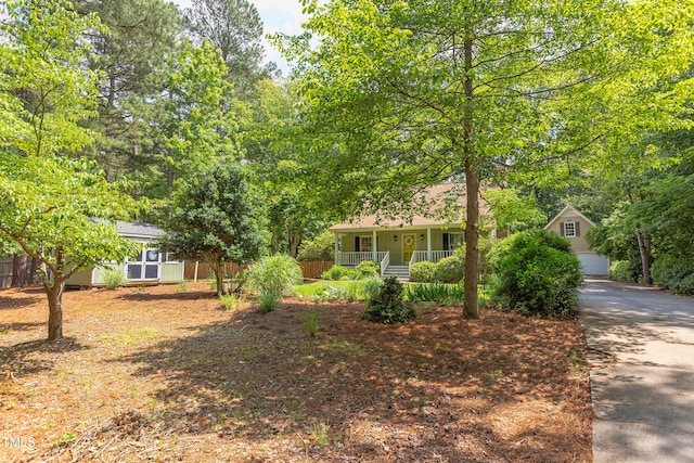 view of property hidden behind natural elements with a porch, fence, a garage, an outdoor structure, and driveway