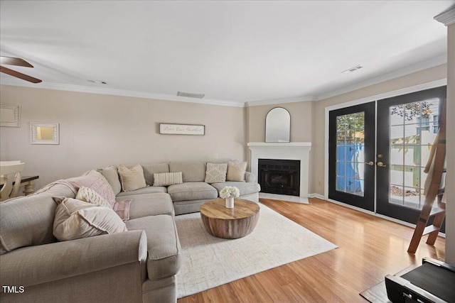 living room featuring ceiling fan, light wood finished floors, crown molding, a fireplace with raised hearth, and french doors
