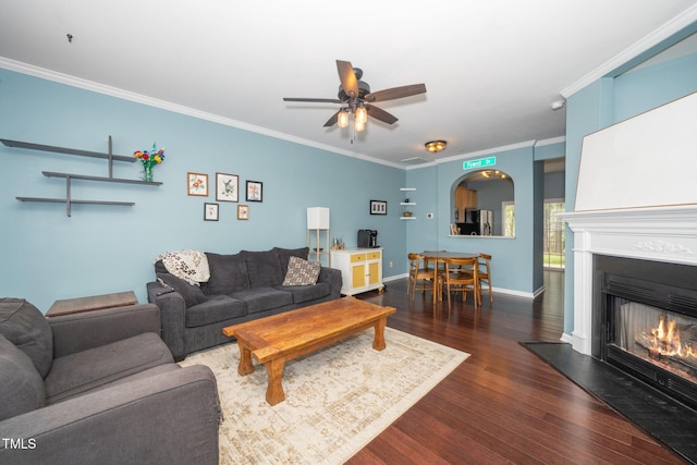 living room with ornamental molding, wood finished floors, a warm lit fireplace, a ceiling fan, and arched walkways