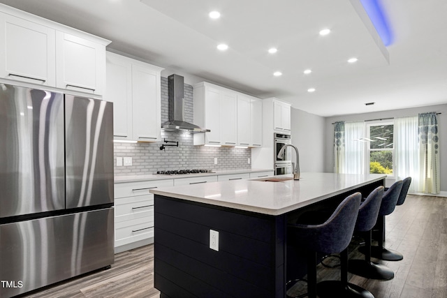 1003 E Sterlington Pl, Apex, NC 27502 - kitchen featuring appliances with stainless steel finishes, wall chimney range hood, a kitchen island with sink, tasteful backsplash, and light wood-style floors