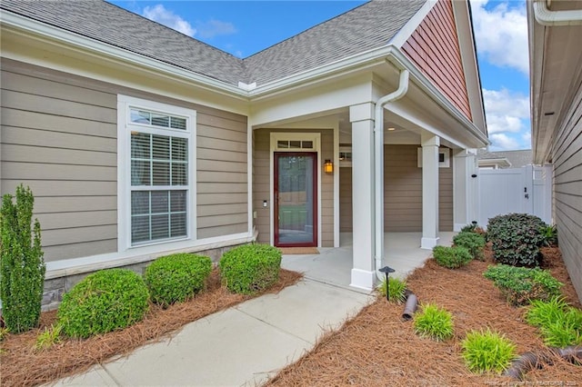 view of exterior entry with a shingled roof, fence, and covered porch