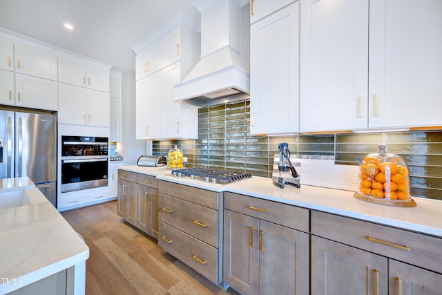 kitchen featuring premium range hood, stainless steel appliances, wood finished floors, backsplash, and white cabinets