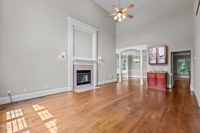 unfurnished living room featuring ceiling fan, a tile fireplace, wood finished floors, a towering ceiling, and ornate columns
