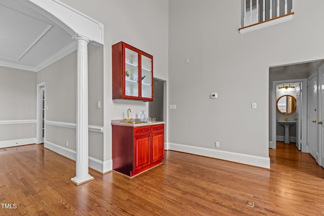 indoor wet bar with ornate columns, wood finished floors, a towering ceiling, baseboards, and crown molding