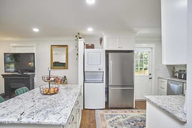 kitchen with stainless steel appliances, stacked washing machine and dryer, white cabinetry, light stone countertops, and wood finished floors