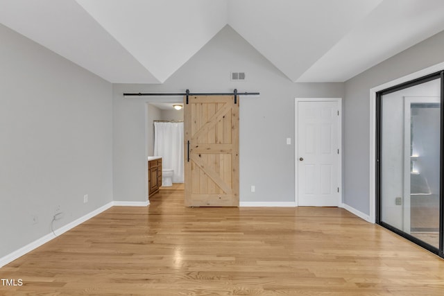 unfurnished bedroom featuring a barn door, light wood-style flooring, vaulted ceiling, baseboards, and connected bathroom