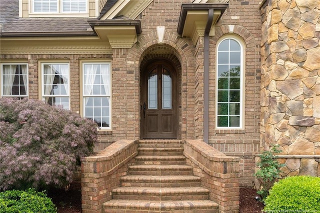  - entrance to property featuring a shingled roof and brick siding