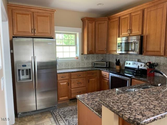 49412 River Run Rd, Albemarle, NC 28001 - kitchen with appliances with stainless steel finishes, a sink, brown cabinetry, dark stone counters, and backsplash
