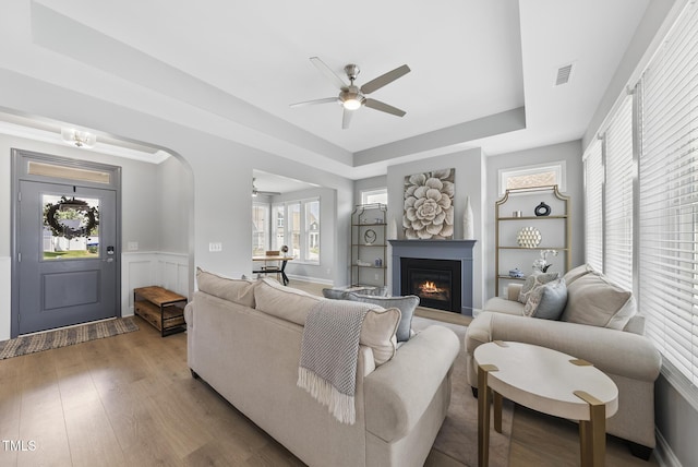 1727 Pinecliff Ct, Fuquay Varina, NC 27526 - living room featuring a ceiling fan, a tray ceiling, a glass covered fireplace, wainscoting, and healthy amount of natural light