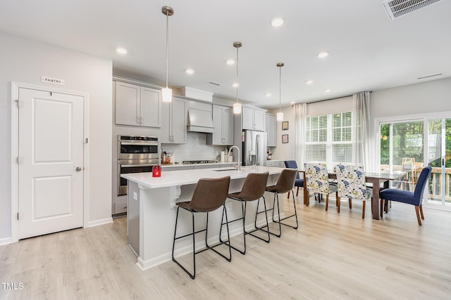 2866 Hunter Woods Dr, Apex, NC 27502 - kitchen featuring appliances with stainless steel finishes, custom range hood, light countertops, a kitchen island with sink, and light wood finished floors