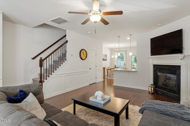 7879 Allscott Way, Raleigh, NC 27612 - living area with stairs, ceiling fan, dark wood-type flooring, a glass covered fireplace, and a wainscoted wall
