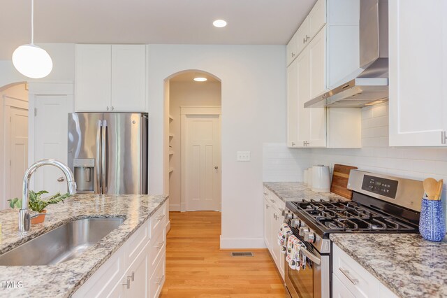 119 W Piedmont Ave, Durham, NC 27707 - kitchen featuring appliances with stainless steel finishes, wall chimney exhaust hood, arched walkways, white cabinetry, and light wood finished floors