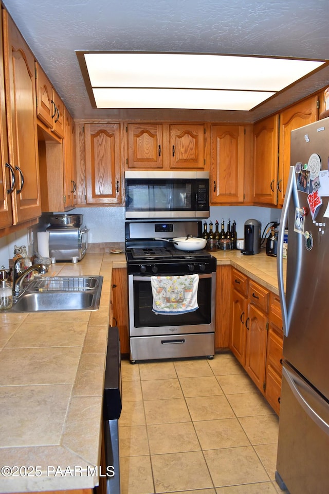 kitchen with tile countertops, light tile patterned floors, brown cabinetry, stainless steel appliances, and a sink