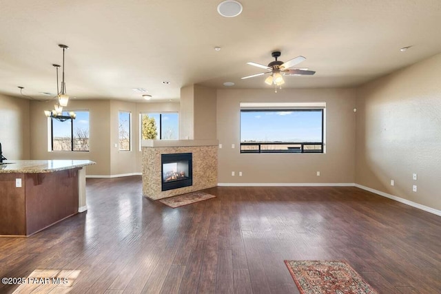 unfurnished living room with dark wood-style flooring, a multi sided fireplace, baseboards, and ceiling fan with notable chandelier