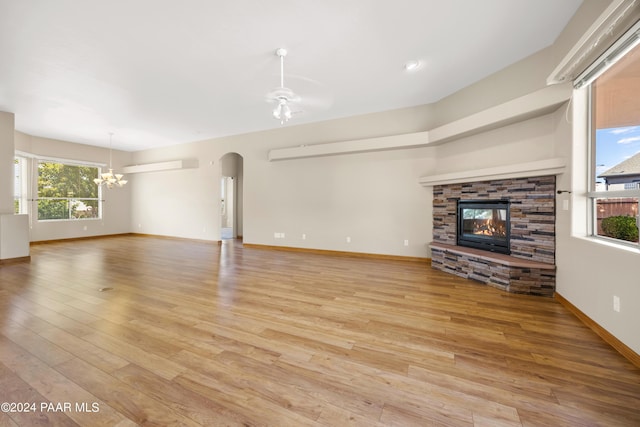 unfurnished living room with ceiling fan with notable chandelier, light wood-type flooring, a stone fireplace, and a healthy amount of sunlight