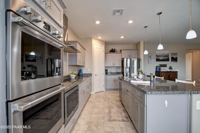 kitchen featuring visible vents, a kitchen island with sink, gray cabinets, stainless steel appliances, and decorative light fixtures