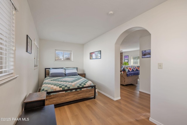 bedroom with multiple windows, light wood-style floors, arched walkways, and baseboards