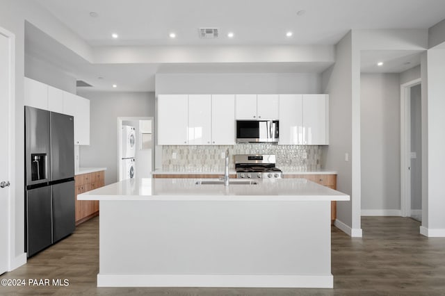 kitchen featuring appliances with stainless steel finishes, dark wood-type flooring, stacked washer / dryer, white cabinetry, and an island with sink