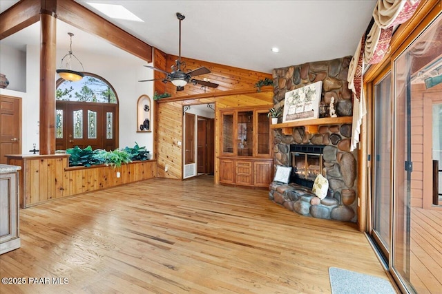 unfurnished living room with vaulted ceiling with skylight, light wood-style floors, a stone fireplace, wood walls, and ceiling fan