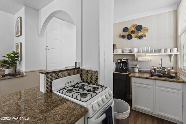 kitchen featuring dark wood-type flooring, white range with gas stovetop, dark stone countertops, arched walkways, and a sink