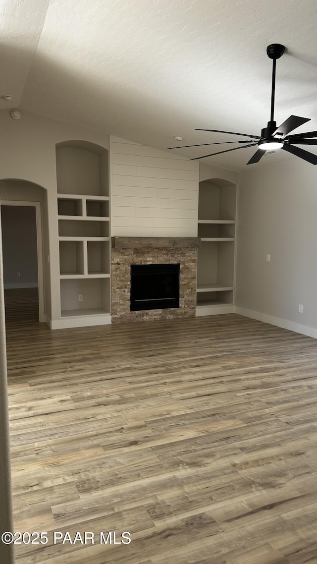 unfurnished living room featuring a textured ceiling, hardwood / wood-style floors, built in features, ceiling fan, and a stone fireplace