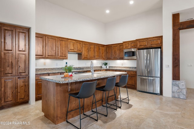 kitchen with a high ceiling, a center island with sink, sink, light stone countertops, and appliances with stainless steel finishes