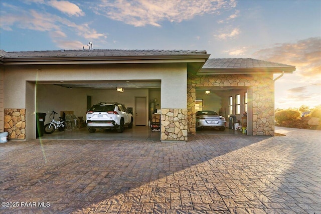 property exterior at dusk with an attached garage, stucco siding, decorative driveway, and stone siding