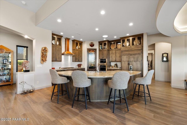 kitchen with appliances with stainless steel finishes, arched walkways, brown cabinetry, wall chimney exhaust hood, and a spacious island