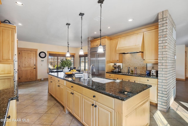 kitchen with stainless steel built in refrigerator, premium range hood, dark stone countertops, decorative light fixtures, and a center island with sink