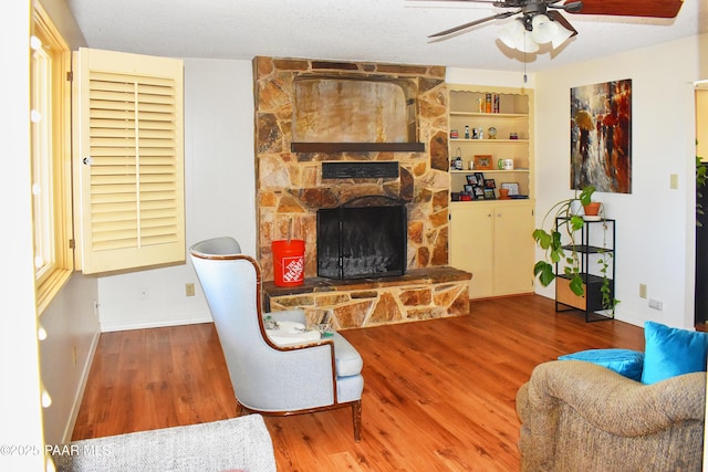 living area featuring a stone fireplace, wood finished floors, a ceiling fan, and baseboards