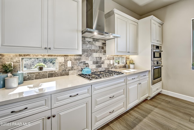 kitchen with wall chimney range hood, appliances with stainless steel finishes, decorative backsplash, white cabinets, and light wood-type flooring