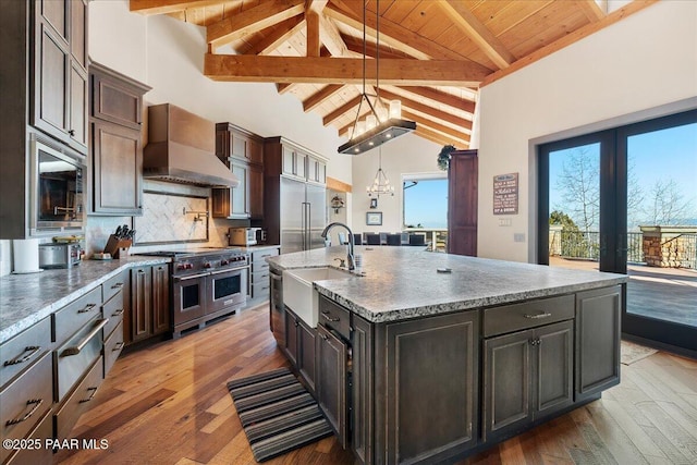 kitchen featuring dark brown cabinetry, custom exhaust hood, built in appliances, decorative light fixtures, and an island with sink