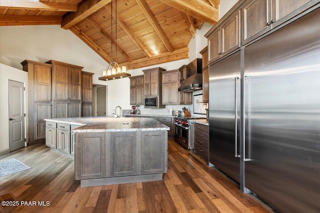 kitchen with decorative light fixtures, wall chimney range hood, built in appliances, light stone countertops, and a center island with sink