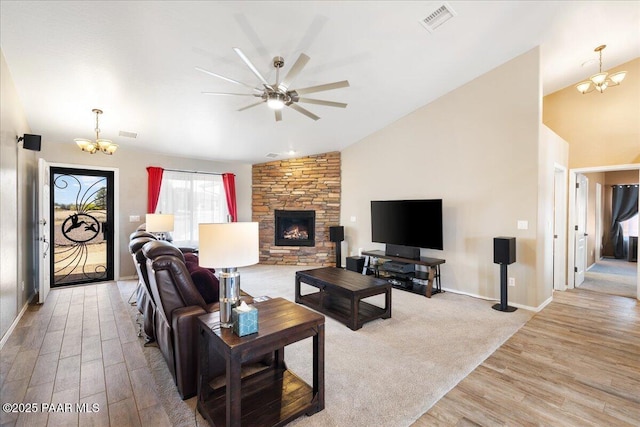 living room with ceiling fan with notable chandelier, vaulted ceiling, light wood-style flooring, baseboards, and a stone fireplace