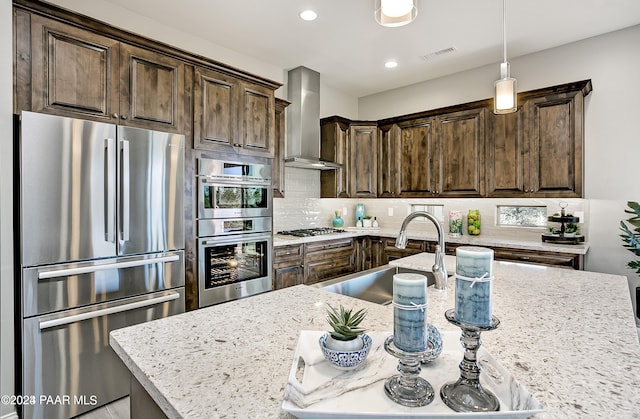 kitchen featuring pendant lighting, wall chimney range hood, sink, stainless steel appliances, and light stone countertops