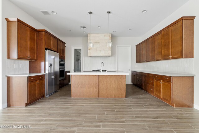 kitchen featuring stainless steel appliances, a kitchen island with sink, sink, decorative light fixtures, and light hardwood / wood-style flooring