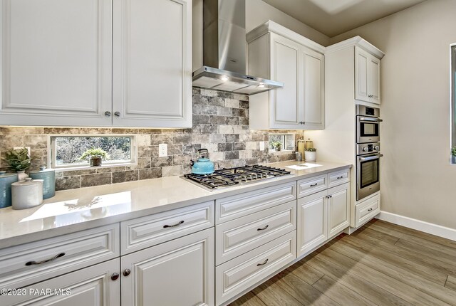 kitchen with decorative backsplash, light countertops, light wood finished floors, stainless steel appliances, and wall chimney exhaust hood