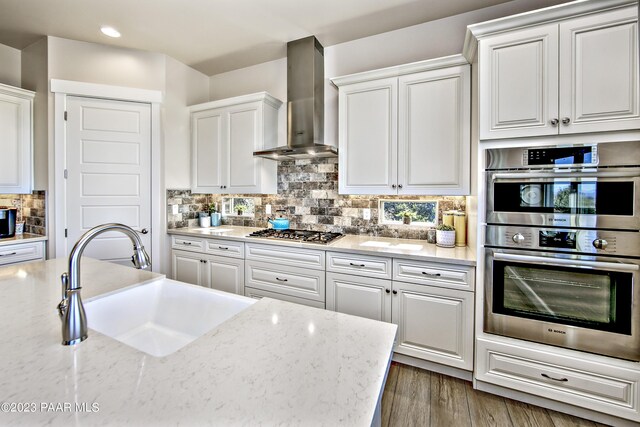 kitchen with wall chimney exhaust hood, appliances with stainless steel finishes, white cabinetry, a sink, and backsplash