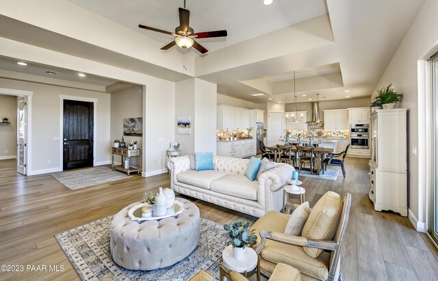 living room with a raised ceiling, ceiling fan with notable chandelier, baseboards, and light wood-type flooring