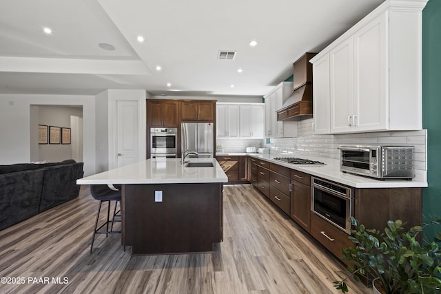 kitchen with visible vents, light wood finished floors, premium range hood, decorative backsplash, and appliances with stainless steel finishes