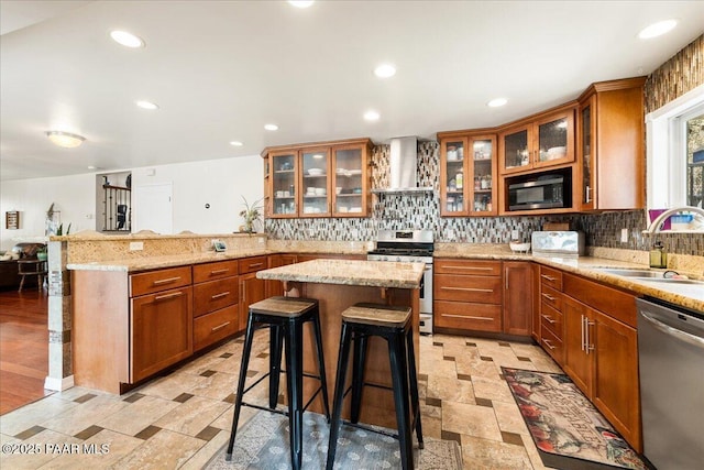 kitchen with wall chimney range hood, sink, a breakfast bar, appliances with stainless steel finishes, and a kitchen island